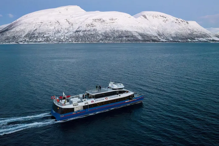 Un catamarano veloce bianco e blu che attraversa un fiordo scuro sotto una montagna innevata