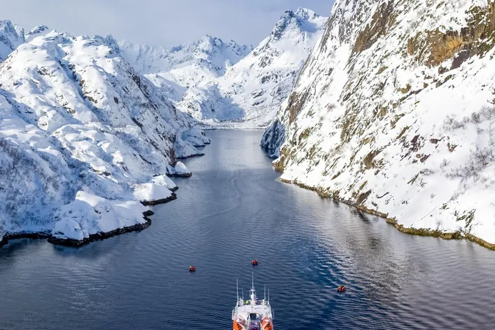 A small expedition ship in a narrow fjord between steep snow-covered mountains
